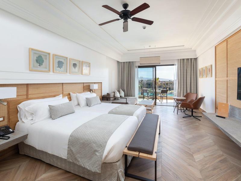 Geräumiges Hotelzimmer mit Doppelbett, Parkettboden, Deckenventilator und Balkon mit Meerblick.