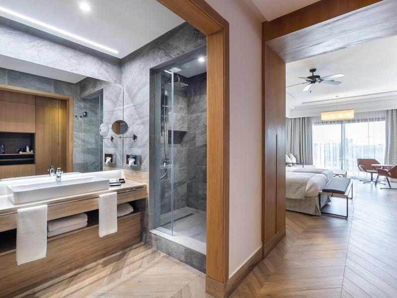 Moderner Hotelzimmerbereich mit Bad, Dusche, Doppelwaschbecken und angrenzendem Schlafzimmer mit Balkon.