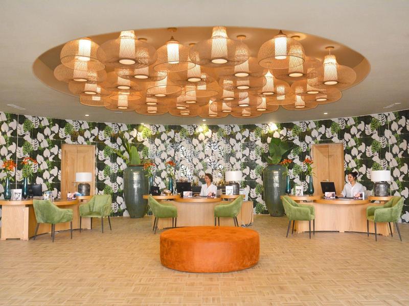 Elegante Empfangshalle mit floralem Wandmuster, grünen Sesseln, runden Holztischen und moderner Deckenleuchte.