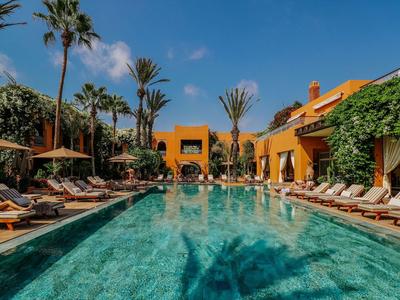 Large pool surrounded by sun loungers, palm trees, and yellow buildings under a blue sky.