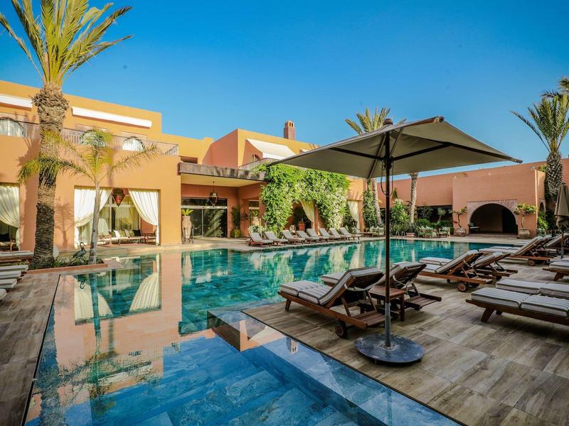 Moderner Außenpool mit Liegestühlen, Sonnenschirm und Palmen vor rosa Gebäude unter blauem Himmel.