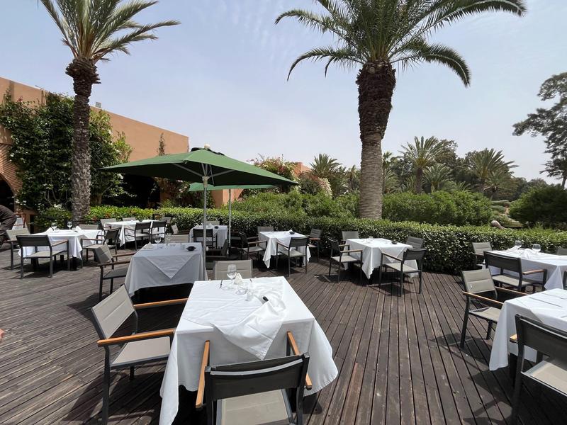 Outdoor-Restaurant mit weißen Tischdecken, Holzboden, grünen Pflanzen und Palmen bei klarem Himmel.