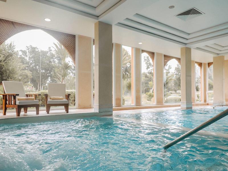 Innenbereich mit Schwimmbecken, großen Bogenfenstern und Sitzbereich mit Stühlen und Tisch.