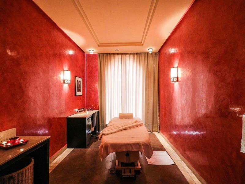 Ein schmaler Massageraum mit roten Wänden, Massagebank in der Mitte und weichem Licht.