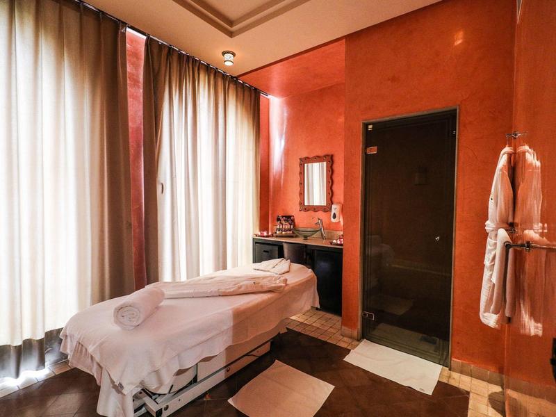 Behandlungsraum mit Massageliege, orangefarbenen Wänden, Vorhängen und Bademänteln an der Wand.