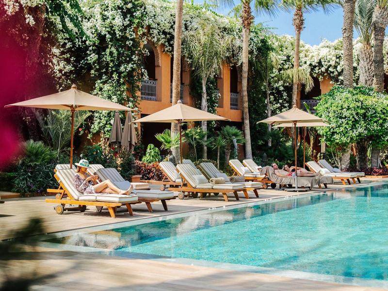 Pool mit blauen Wasser, Liegestühlen und Sonnenschirmen, umgeben von hohen Palmen und grünen Pflanzen.