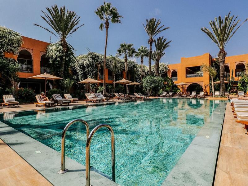 Großer Pool mit kristallklarem Wasser, umgeben von Palmen und orangefarbenen Gebäuden.