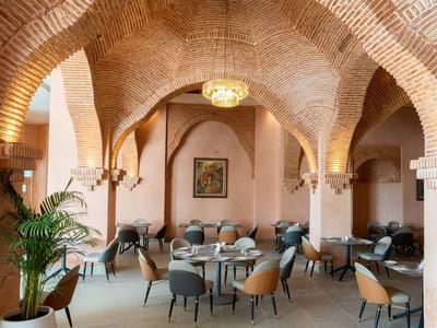 Gemütlicher Gastraum mit Backsteinbögen, runden Tischen, gepolsterten Stühlen und Pflanzen.