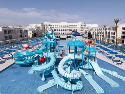 Parque acuático con varios toboganes y juegos de agua frente a un hotel con muchas tumbonas azules.
