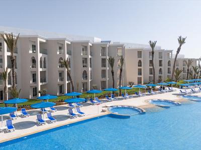 Gran piscina al aire libre con sillas azules y edificios modernos del hotel al fondo.