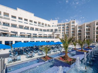 Modernes, weißes Hotel mit mehreren Etagen, blauen Sonnenschirmen und Palmen um einen Wasserbrunnen.