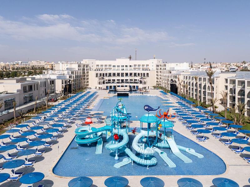 Großer Außenpool mit Wasserrutschen und vielen blauen Liegestühlen vor weißen Hotelgebäuden.