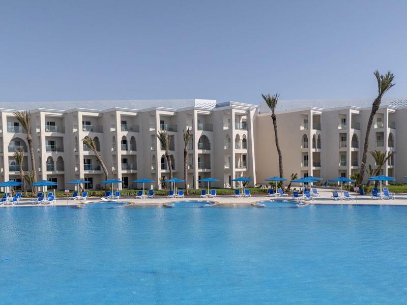 Großer blauer Pool vor vierstöckigem weißem Hotel mit Palmen und blauen Liegen.