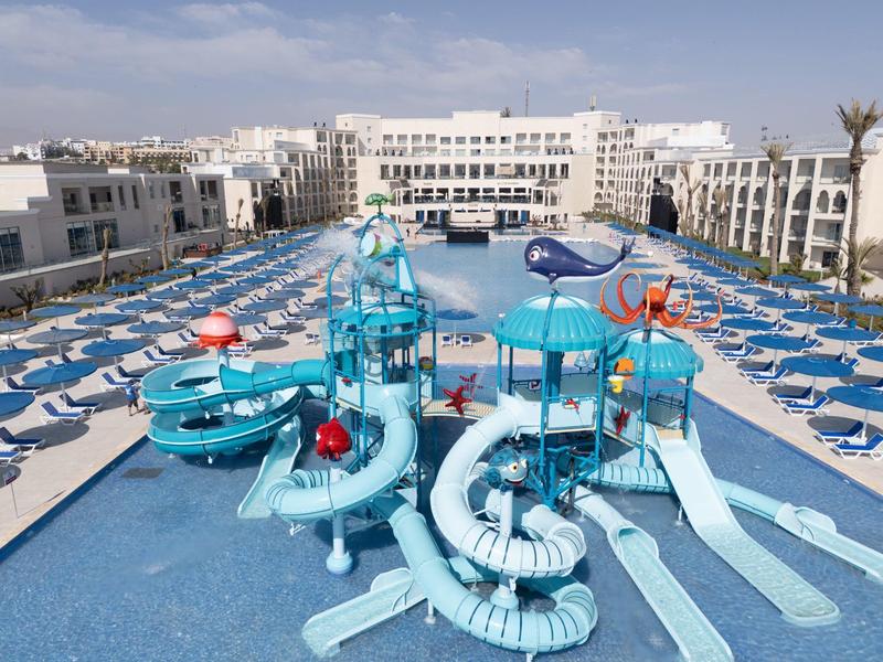 Großes Schwimmbad mit blauen Wasserrutschen und vielen blauen Liegestühlen rundherum.
