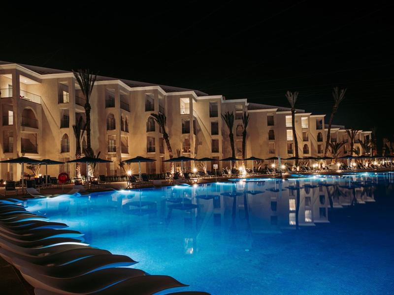 Großer beleuchteter Hotelpool bei Nacht, umgeben von mehrstöckigen Gebäuden und Palmen.