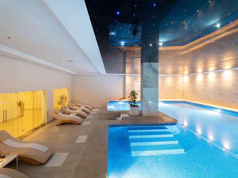 Moderner Indoor-Pool mit blau beleuchtetem Wasser, Liegestühlen und heller Wandbeleuchtung.