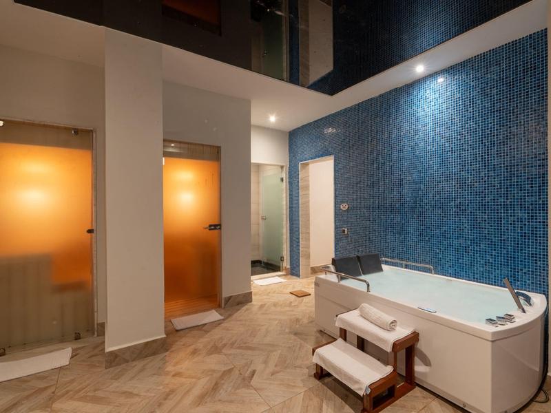 Moderner Wellnessbereich mit blauer Mosaikwand, Wanne, zwei Duschen und hellen Holzbodenfliesen.