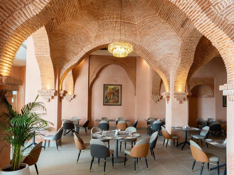 Gemütlicher Gastraum mit Backsteinbögen, runden Tischen, gepolsterten Stühlen und Pflanzen.