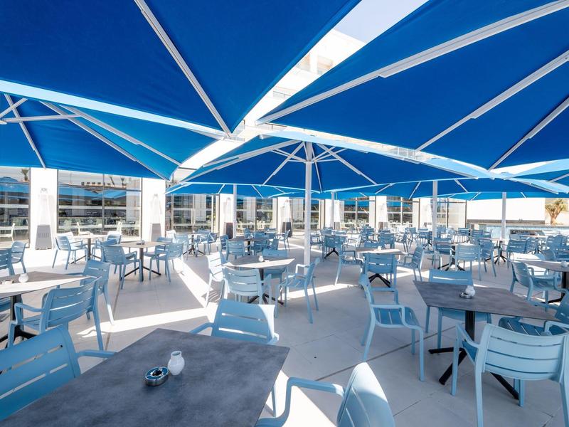 Große, helle Außenterrasse mit blauen Sonnenschirmen und weißen Stühlen an Tischgruppen.
