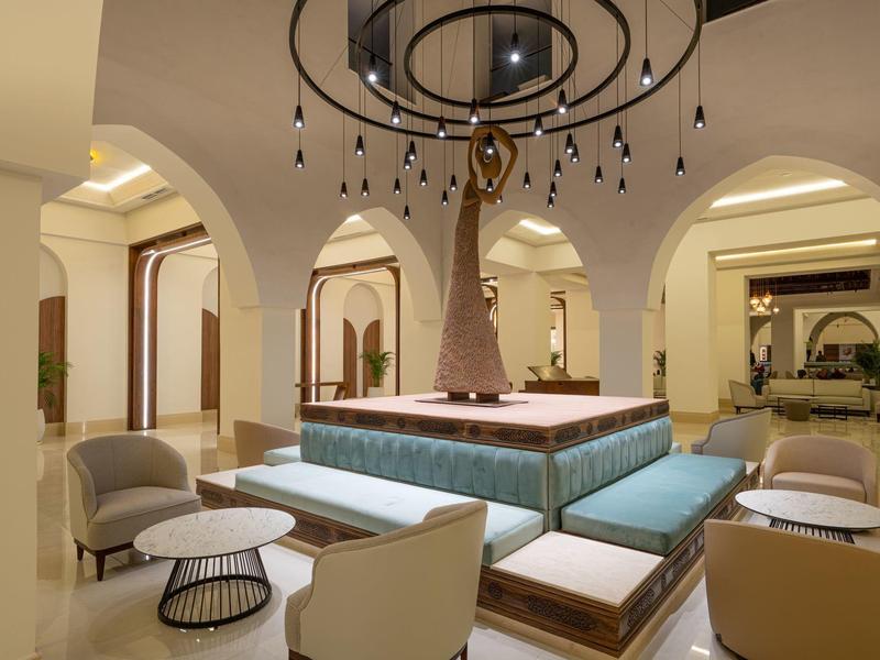 Moderner Loungebereich mit zentralem Kunstbaum, Sitzbänken, Sesseln und runder Lampe in warmem Licht.