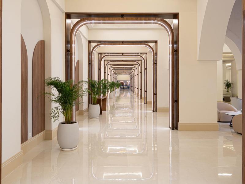 Langer, heller Gang mit weißen Fliesen und großen Pflanzen in Töpfen entlang der Wände.