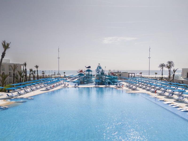 Großer blauer Pool mit vielen Liegestühlen, Wasserrutschen und Palmen am Rand unter hellem Himmel.