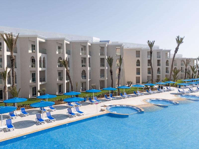 Großer blauer Pool mit vielen blauen Liegestühlen vor weißem Hotelgebäude und Palmen.