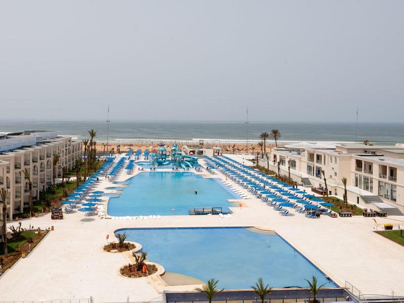 Großer rechteckiger Pool zwischen zwei langen Gebäuden mit Blick aufs Meer im Hintergrund.