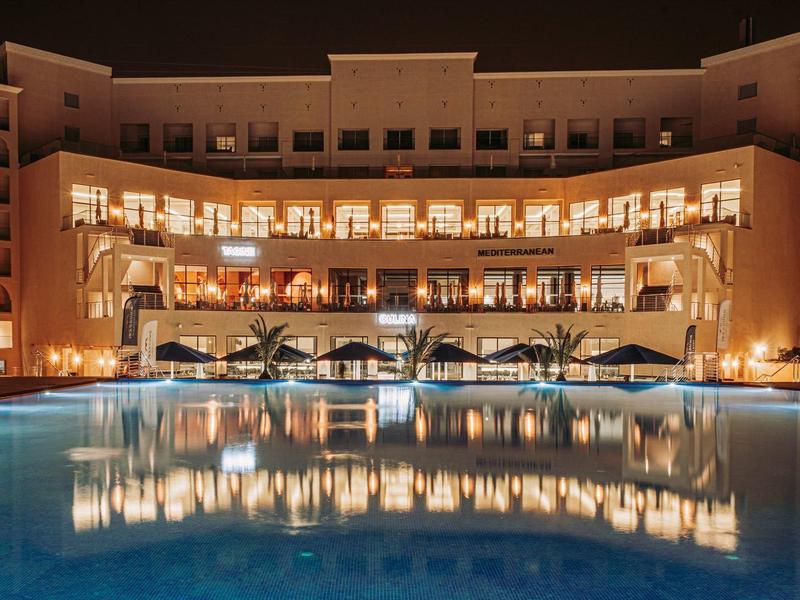 Großes Hotel mit warmen Lichtern und beleuchtetem Pool bei Nacht, spiegelglatte Wasseroberfläche.