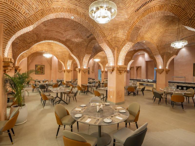 Restaurant mit Backsteinbögen, runden Tischen, beige Stühlen und warmem Licht.