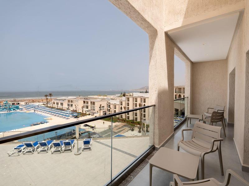 Balkon mit Stühlen und Glasgeländer, Blick auf Pool, Liegestühle und Häuser bei klarem Himmel.