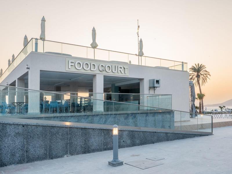 Moderner Food-Court mit Glasgeländer, weißer Fassade und Palme bei Sonnenuntergang.