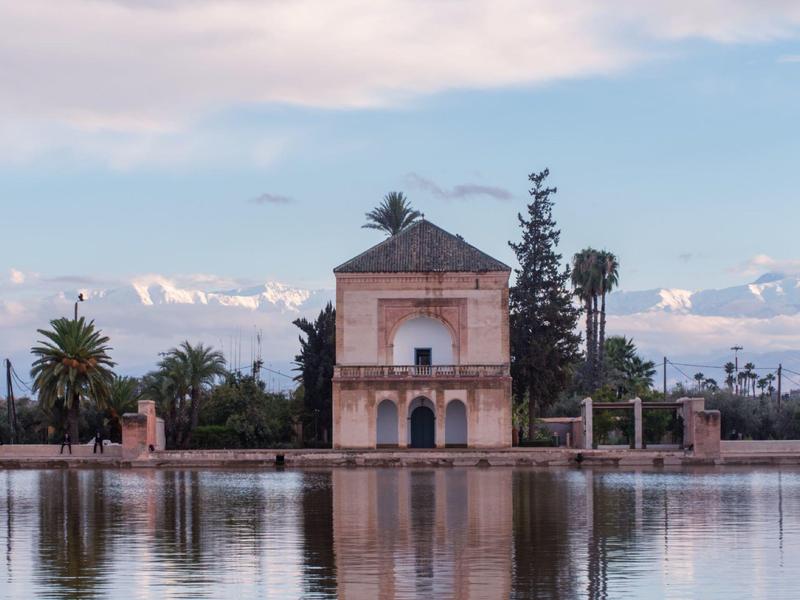 Ein altes Gebäude mit spitzem Dach steht an einem ruhigen Wasserbecken, umgeben von Palmen.