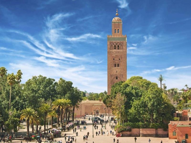 Hoher Turm aus roten Steinen, umgeben von grünen Bäumen und einem belebten Platz darunter.