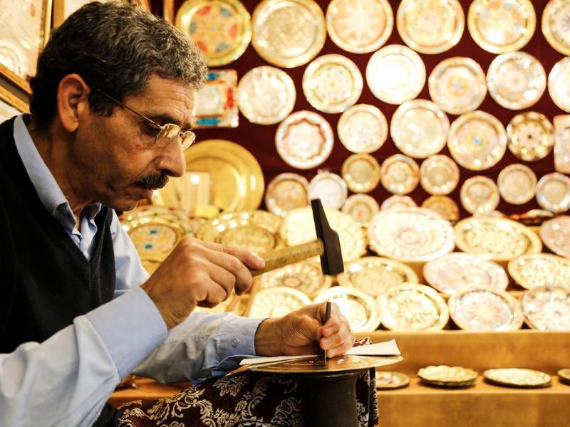 Ein Mann mit Brille schlägt mit einem kleinen Hammer auf ein Metallobjekt in einer Werkstatt mit vielen runden Platten.