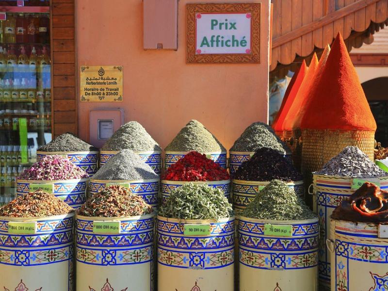 Bunte Gewürzbehälter mit verschiedenen Kräutern und Gewürzen vor einer orangen Wand.