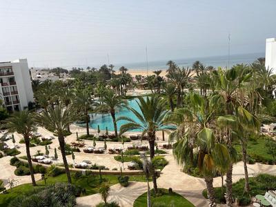Hotel complex with pool, palm trees, and sea view on a sunny day.