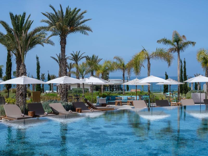 Luxe zwembadomgeving met ligbedden, witte parasols en palmbomen onder een heldere hemel.