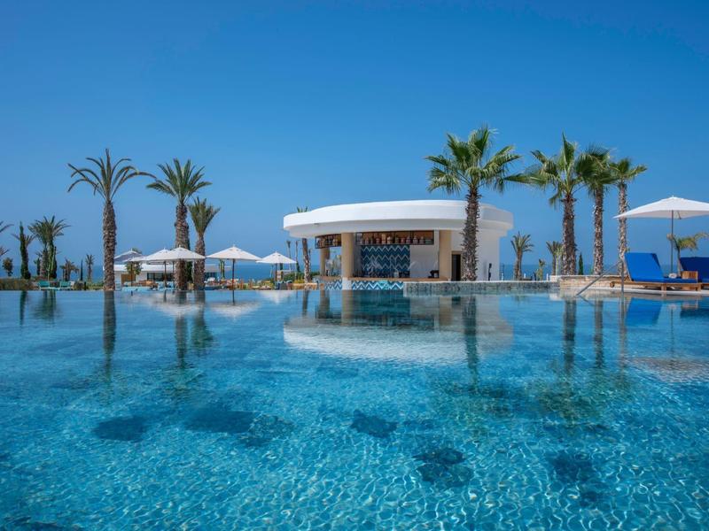 Een zwembad met helder water omgeven door parasols en palmbomen onder een blauwe lucht.