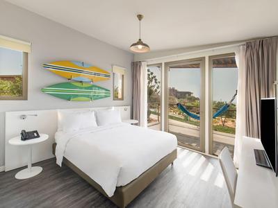 Bright hotel room with double bed, surfboards on the wall, and view of pool and sea.