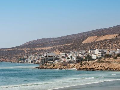 Coastal town with beach, waves, and mountains in the background under a clear sky.