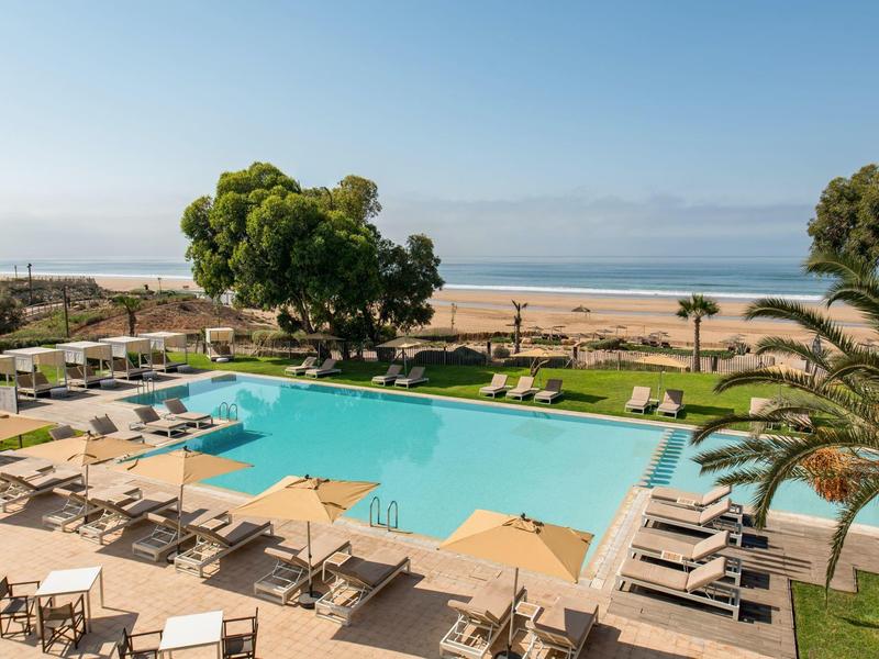 Grande piscine avec chaises longues et parasols face à la plage et la mer.