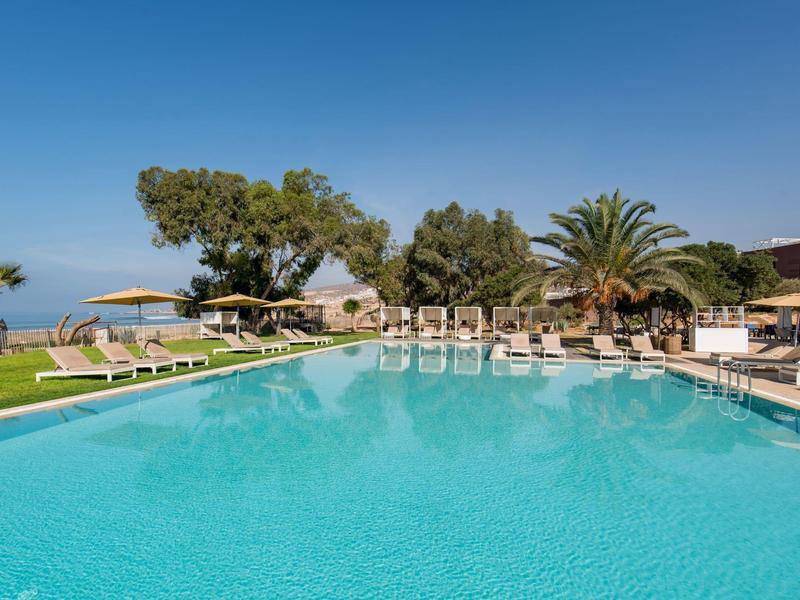 Grand piscine avec chaises longues et parasols, entourée de palmiers sous un ciel bleu clair.