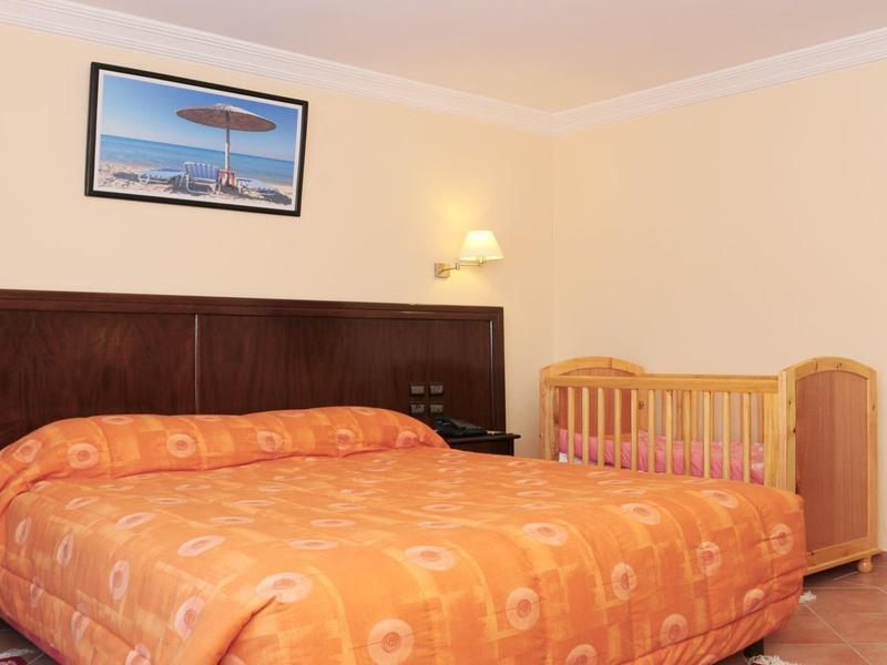 Schlafzimmer mit großem Doppelbett, orangefarbener Decke, Kinderbett und Wandlampe an beiger Wand.