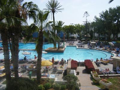 Duży basen hotelowy z palmami, leżakami i parasolami w słoneczny dzień.