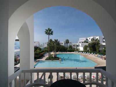 Widok przez łuk na zewnętrzny basen z palmami i białymi budynkami hotelowymi pod czystym niebem