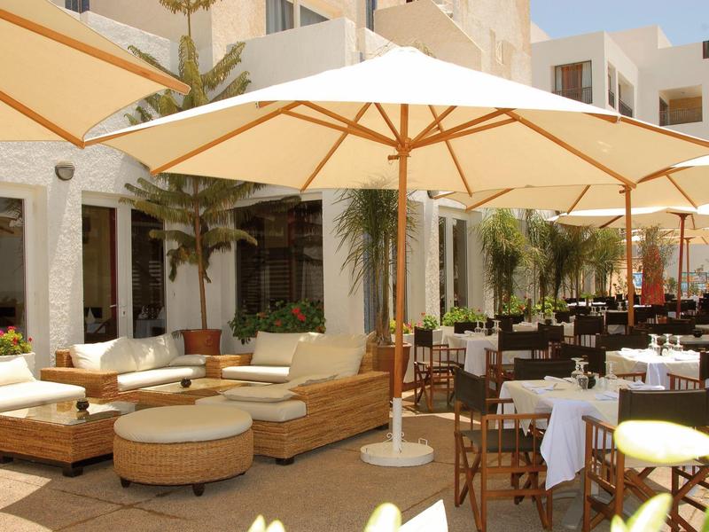 Freiluftrestaurant mit weißen Sonnenschirmen, Rattanmöbeln und Holztischen vor einem hellen Gebäude.