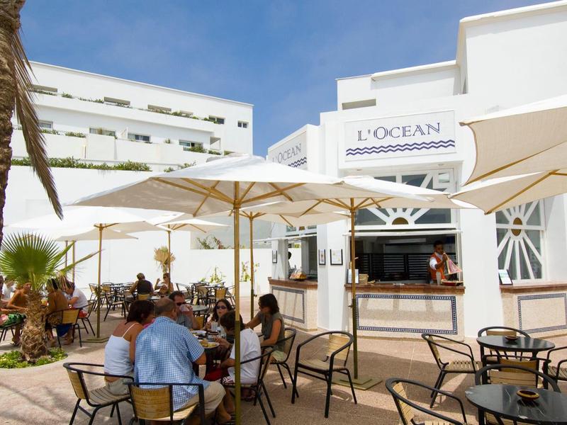 Terrasse eines Cafés mit weißen Sonnenschirmen, braunen Stühlen und Palmen bei sonnigem Himmel.