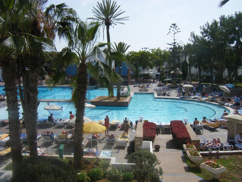Großer Pool mit Palmen, vielen Liegestühlen, Sonnenschirmen und Menschen bei sonnigem Wetter.