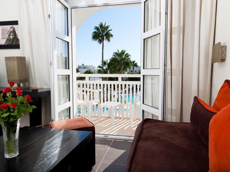 Wohnzimmer mit brauem Sofa, orangen Kissen, Vase mit Rosen und Blick auf Terrasse mit Palme.